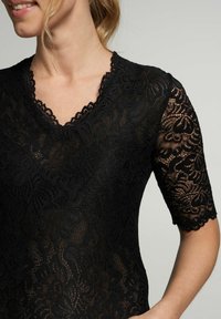 Schwarzes Spitzenoberteil mit kurzen Ärmeln, das über einen gewellten V-Ausschnitt verfügt. Das filigrane Blumenmuster hebt die zarte Textur des Stoffes hervor.