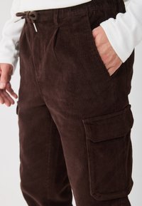 Brązowe spodnie cargo z kordurowej tkaniny o teksturowanej prążkowanej powierzchni, elastycznym pasie i bocznymi kieszeniami. Cechy projektu obejmują luźny krój oraz dodatkową kieszeń cargo.