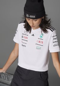 Camisa polo de manga corta blanca con detalles en negro y varios logotipos de patrocinadores. Combinada con pantalones negros, el conjunto incluye un gorro de punto negro.
