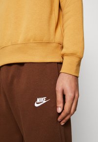 Keltainen collegepaita resoratuilla hihoilla, yhdistettynä ruskeisiin collegehousuihin, joissa valkoinen Nike-logo. Lähikuva kankaan koostumuksesta ja ompeleista.