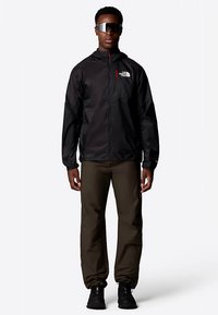 Crna lagana jakna s logom, zatvaranje na patentni zatvarač, duži rukavi, u kombinaciji s maslinastozelenim hlačama i crnim cipelama, nosi osoba.