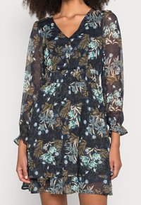 Robe florale avec un décolleté en V profond, des manches longues en voile, une taille froncée et une jupe à volants dans des motifs floraux bleu foncé, vert et beige.