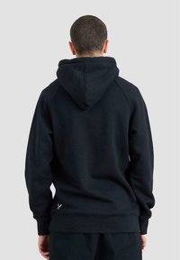 Mężczyzna w czarnej bluzie z kapturem widziany od tyłu na jednolitym jasnoszarym tle.