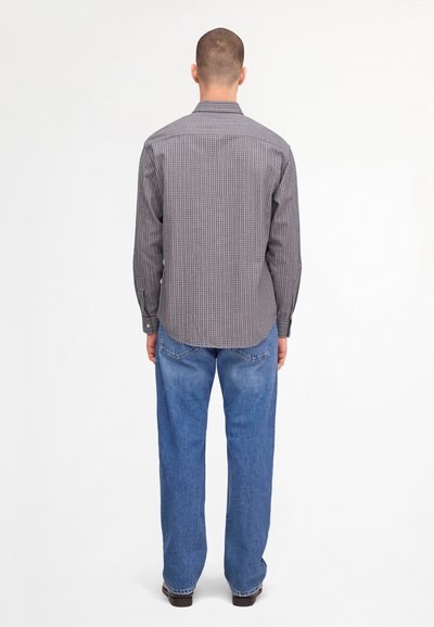 Homme portant une chemise à manches longues à carreaux et un jean bleu, debout de dos devant un fond uni clair.