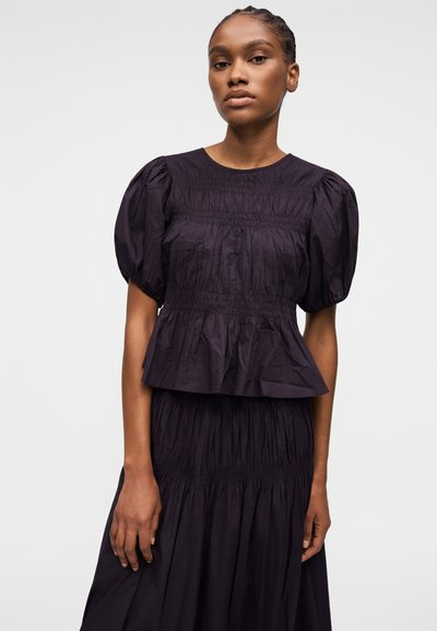 Femme aux cheveux tressés, portant un chemisier noir à manches bouffantes et une jupe noire assortie, debout devant un fond clair uni.