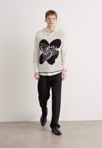 White knit vest featuring a black abstract design, worn over a white button-up shirt, paired with black trousers and black shoes.