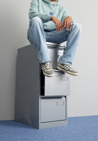 Metallarkivskåp i grått med tre lådor. Person som sitter på toppen i en mintgrön hoodie och ljusblå jeans, med grå sneakers.