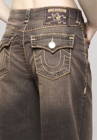 Hnědé džínové kalhoty s kontrastním žlutým šitím, ozdobnými zadními kapsami a štítkem s logem "True Religion" na pasu.