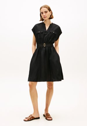 Kvinde iført en sort kortærmet kjole med bælte og forlommer samt brune flade sandaler, stående mod en ensfarvet hvid baggrund.