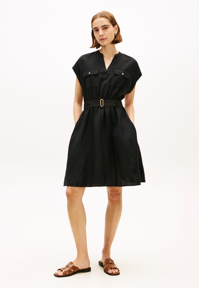 Vrouw die een zwarte jurk met korte mouwen en ceintuur draagt, voorzien van voorzakken, en bruine platte sandalen, staand tegen een effen witte achtergrond.