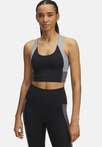 Vrouw in zwart en grijs sportcrop-top en hoge taille leggings, staande tegen een effen lichte achtergrond.