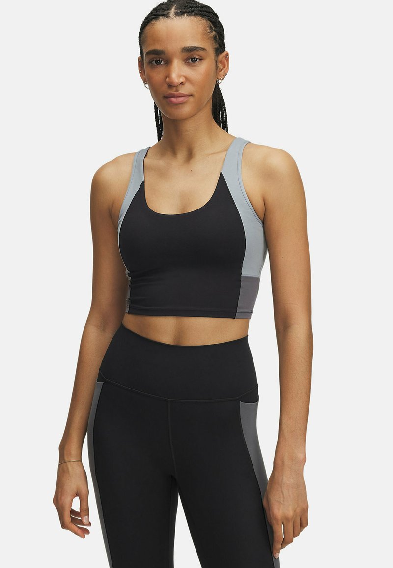 Vrouw in zwart en grijs sportcrop-top en hoge taille leggings, staande tegen een effen lichte achtergrond.