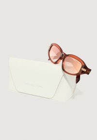 Occhiali da sole oversize marroni con finitura lucida, appoggiati su un astuccio bianco texture con "MICHAEL KORS" impresso.