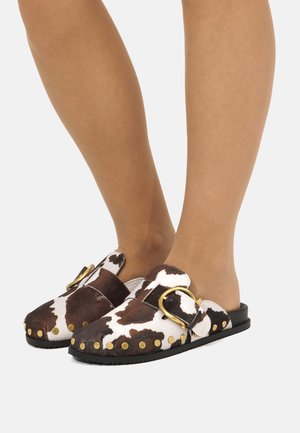 Scarpe slip-on con stampa mucca marrone e bianca, con borchie dorate e grande fibbia dorata, indossate ai piedi su sfondo bianco.