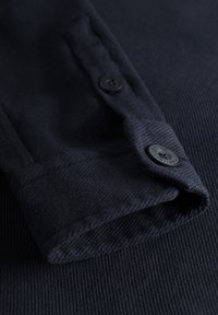 Tessuto blu scuro con texture a righe verticali, dotato di un polsino con due bottoni neri per la chiusura e un bordo pulito e sagomato.