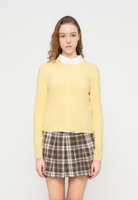 Pull en maille jaune à manches longues, associé à une jupe plissée à carreaux dans des tons marron, blanc et gris. Col blanc visible en dessous.