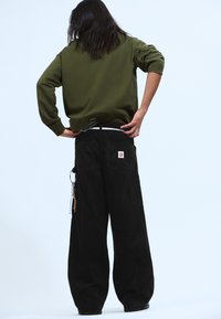 Person mit langen Haaren, der einen olivgrünen Sweatshirt und weite schwarze Hosen trägt, mit dem Rücken zur Kamera, der den Hosenbund mit den Händen im Rücken anpasst.