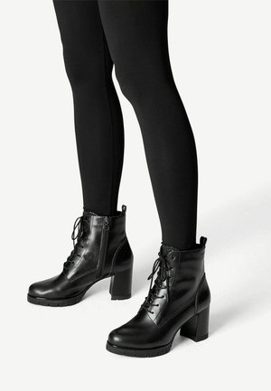 Zwarte leren enkellaarsjes met hak, voorzien van veters aan de voorkant en zijritsen. Met textuuraccenten en een stevige hak. Draagt zwarte leggings.