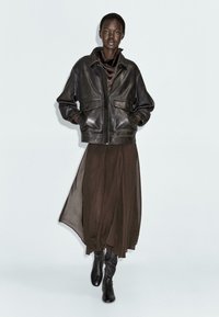 Giacca di pelle nera con tasche frontali, indossata sopra una gonna fluida marrone scuro. L'outfit è completato da stivali neri alti fino al ginocchio.