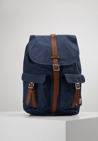 Granatowy plecak z materiału z klapą składana z przodu, brązowe skórzane paski i dwie przednie kieszenie. Posiada srebrne okucia i teksturowaną powierzchnię.