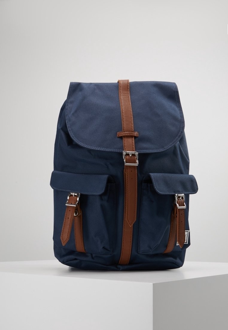 Granatowy plecak z materiału z klapą składana z przodu, brązowe skórzane paski i dwie przednie kieszenie. Posiada srebrne okucia i teksturowaną powierzchnię.