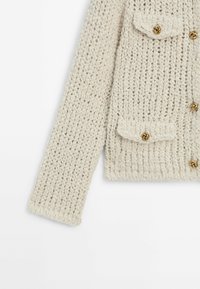 Manche et torse d'un cardigan en maille épaisse crème avec des boutons en forme de nœud doré et des poches à rabat sur fond blanc.
