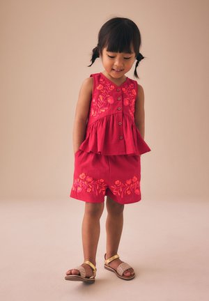 Jeune fille portant un haut sans manches rouge et un short avec une broderie florale, debout et regardant vers le bas, les mains dans les poches.