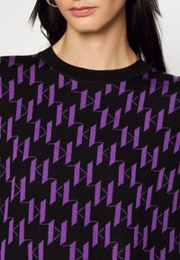 Maglione a maglia con base nera che presenta un motivo geometrico viola ripetuto. Collo rotondo con una texture liscia. Nessun altro dettaglio visibile.