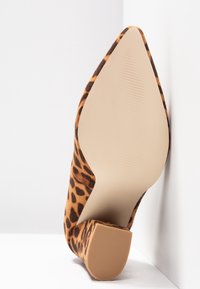Chaussure à bout pointu imprimée léopard avec talon bloc, montrée appuyée contre un coin blanc avec semelle visible et bout de talon texturé.