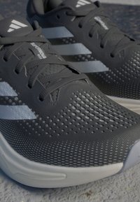 Chaussures de sport gris avec un dessus en maille texturée, un design perforé et une marque à trois bandes blanches. Elles possèdent une semelle rembourrée et des lacets.