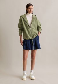 Lichtgroene rits hoodie met een relaxte pasvorm en een trekkoord onderaan, gecombineerd met een donkere denimrok en witte sneakers.
