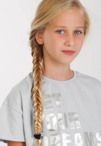 Blond haar in een gevlochten stijl, dragend een grijze T-shirt met "LET YOUR DREAMS" in zilveren tekst. Eenvoudige oorbel zichtbaar. Lichte achtergrond.