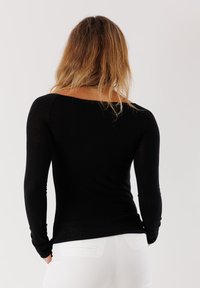 Top nero a maniche lunghe con scollatura a barca, caratterizzato da maniche aderenti e una texture liscia, abbinato a pantaloni bianchi.