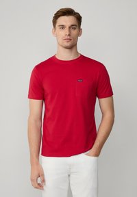 Rode crew-neck t-shirt van zachte katoen, voorzien van een zak op de linkerborst en een klein merkembleem op de zak. Draagt met witte broek.