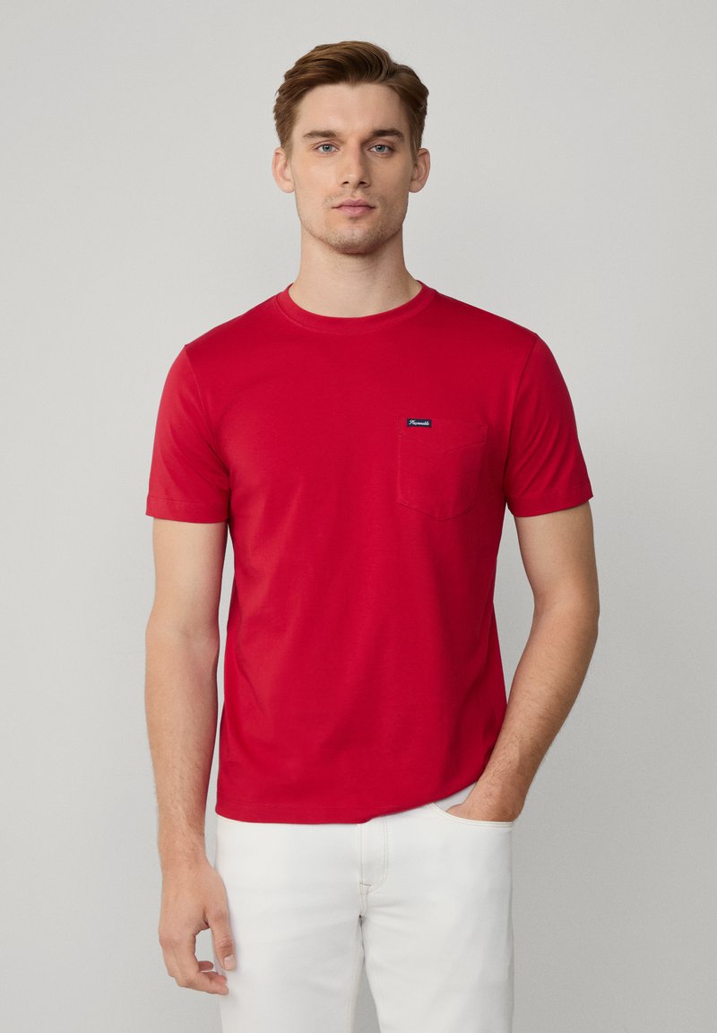 Rode crew-neck t-shirt van zachte katoen, voorzien van een zak op de linkerborst en een klein merkembleem op de zak. Draagt met witte broek.