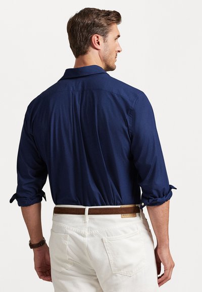Dunkelblaues Hemd mit hochgekrempelten Ärmeln, Button-Down-Kragen und Rückenschlitzen, kombiniert mit hellen Hosen und einem braunen Gürtel.