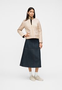 Γυναίκα με μπεζ μπουφάν με καπιτονέ ύφασμα, midi φούστα από σκούρο τζιν, λευκές κάλτσες και μπεζ αθλητικά παπούτσια, στέκεται με το ένα χέρι στη μέση σε απλό φόντο.