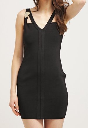 Kobieta ubrana w czarną, bezrękawnikową mini sukienkę z dekoltem w serek i dekoracyjną złotą spinką na lewym ramiączku na jednolitym tle.