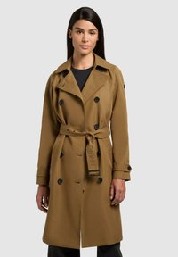 Donna con capelli neri lisci che indossa un trench marrone con cintura sopra una maglietta nera, in piedi contro uno sfondo chiaro e uniforme.