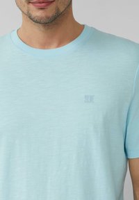 Jasnoniebieska bawełniana koszulka z krótkim rękawem, z okrągłym dekoltem i subtelnym logo w lewym górnym rogu. Gładka faktura.