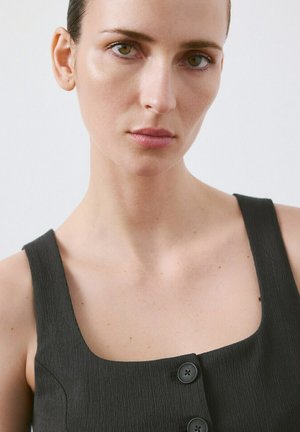 Jonge vrouw met een lichte huid en groene ogen, gekleed in een mouwloos donker gestructureerd topje met grote knopen, neutrale uitdrukking, effen witte achtergrond.