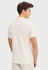 Kremowa bawełniana koszulka polo z klasycznym kołnierzykiem, krótkimi rękawami i nieco dopasowanym krojem, z małym rozcięciem po bokach u dołu.