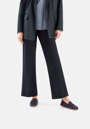 Personne portant un pantalon large noir, un blazer noir, une chemise grise et des mocassins noirs, debout devant un fond blanc.