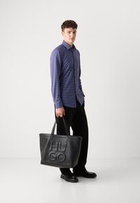 Camisa de botones azul con estampado, bolsa tote de cuero negro con "HUGO" grabado, pantalones de pierna ancha negros y zapatos negros con acentos texturizados.