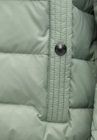 Secção de casaco acolchoado verde claro com um design texturizado e acolchoado. Apresenta um botão de pressão preto e um detalhe de costura proeminente.