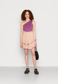 Top roxo de um ombro só combinado com uma saia floral com babados em tons de rosa e amarelo. Sandálias de plataforma pretas e uma bolsa preta texturizada.