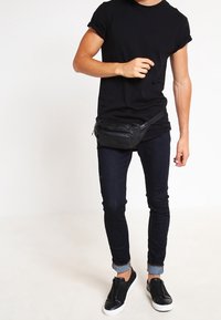 Svart midjeväska med texturerad yta bärs över en åtsittande svart t-shirt, i kombination med mörka jeans och svarta slip-on skor.