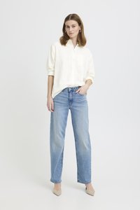 Žena stojící, oblečená v volné bílé košili na knoflíky, světle modrých džínech se rovnými nohavicemi a béžových lodičkách na špičce, na jednobarevném pozadí.