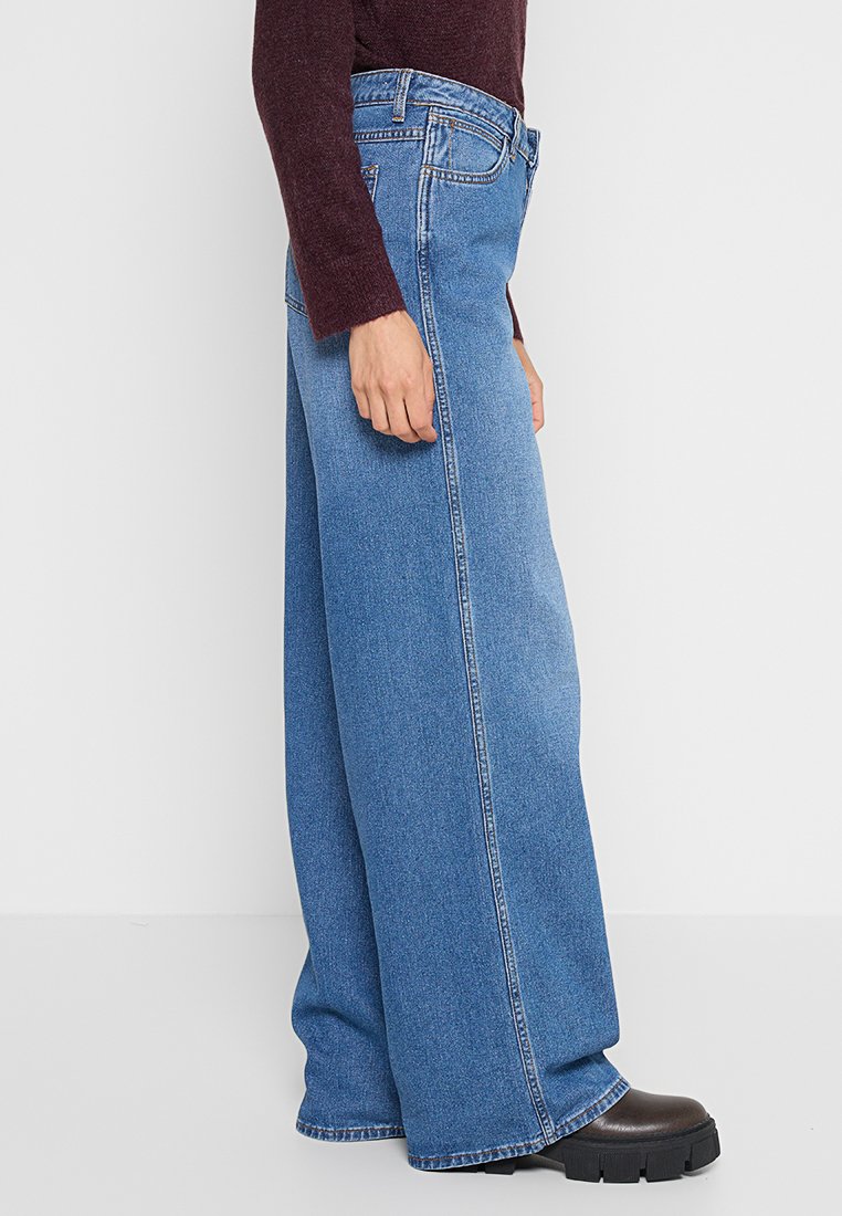 Szerokie niebieskie dżinsy z wysokim stanem, z prostym krojem, klasycznymi szwami i subtelną fakturą. Widać czarne platformy.