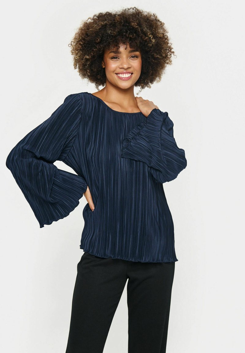 Tmavomodrá skladaná blúzka so širokým výstrihom a rozšírenými rukávmi, s hladkou textúrou a jemným leskom. Skombinovaná s čiernymi nohavicami.